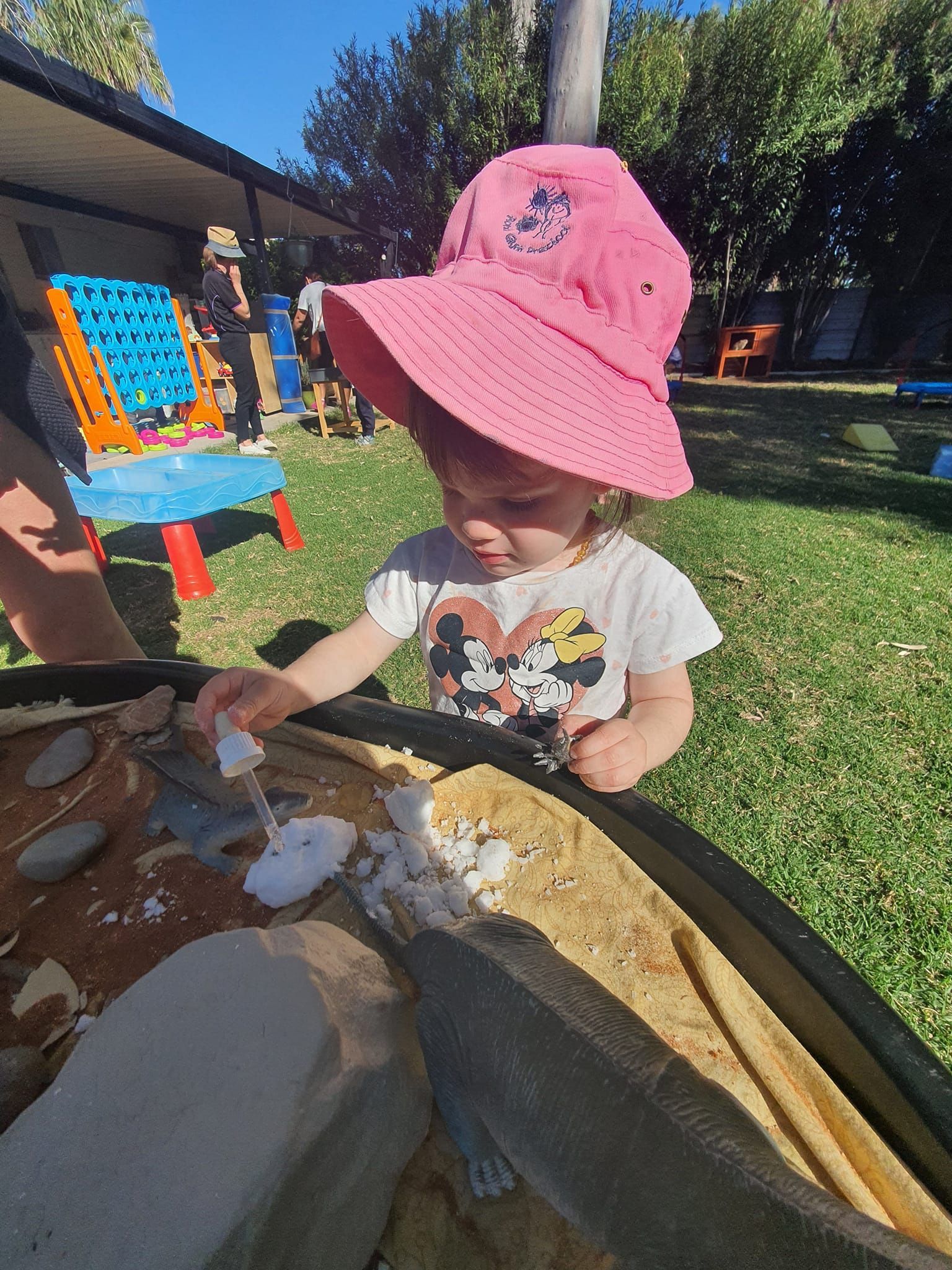 Girl in pink hat plays with materials in a sandbox, outdoors on a sunny day.