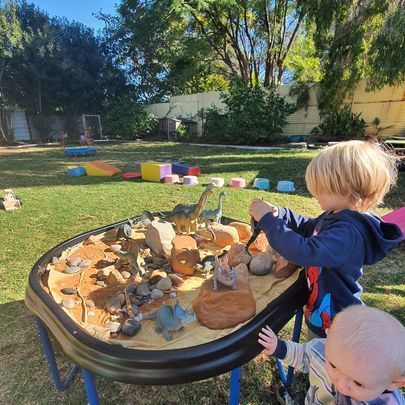 Two young children playing with dinosaur toys in a sandbox outdoors.