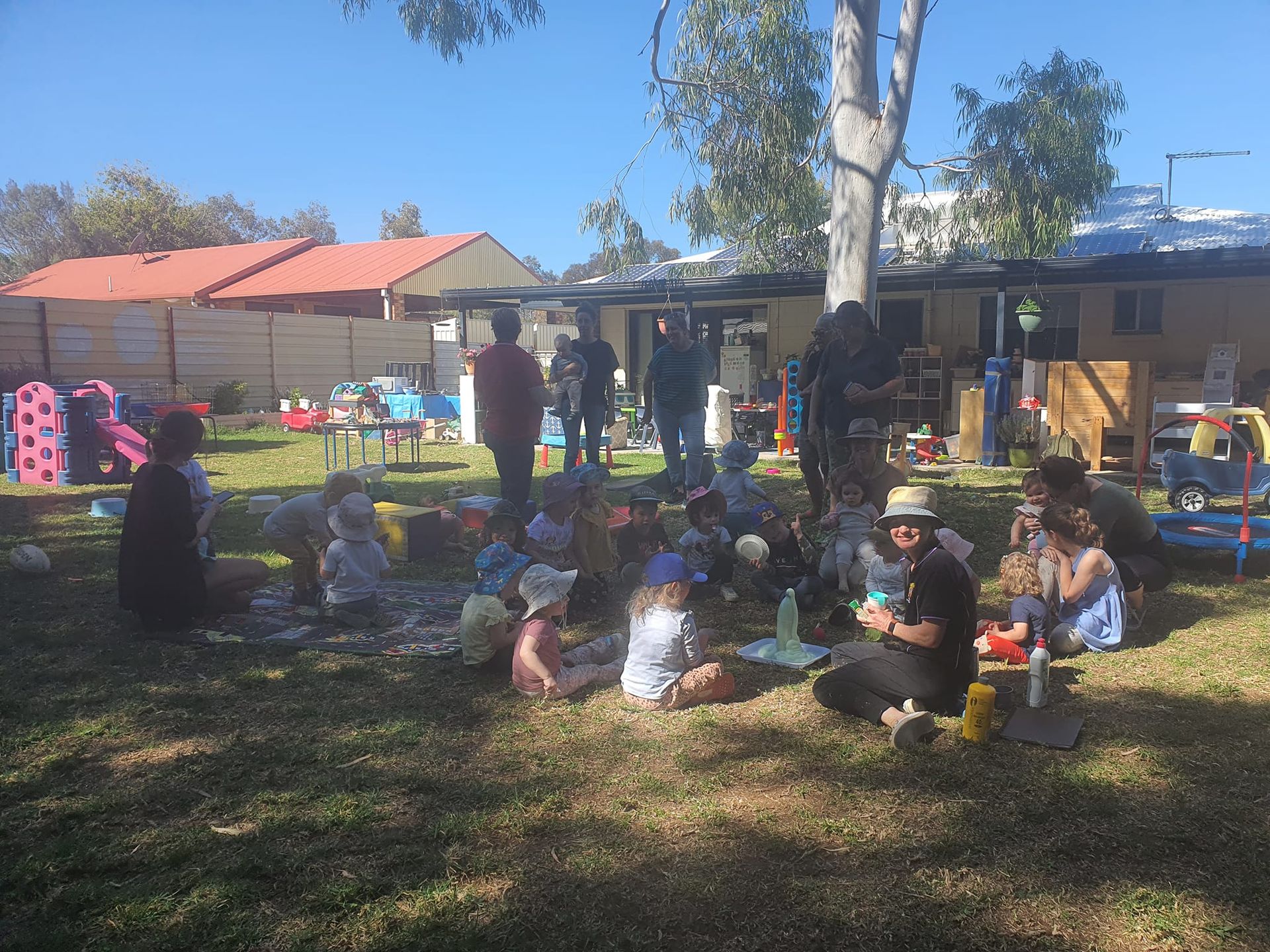 Children and adults gather outdoors, some seated on grass, near a building, toys scattered.