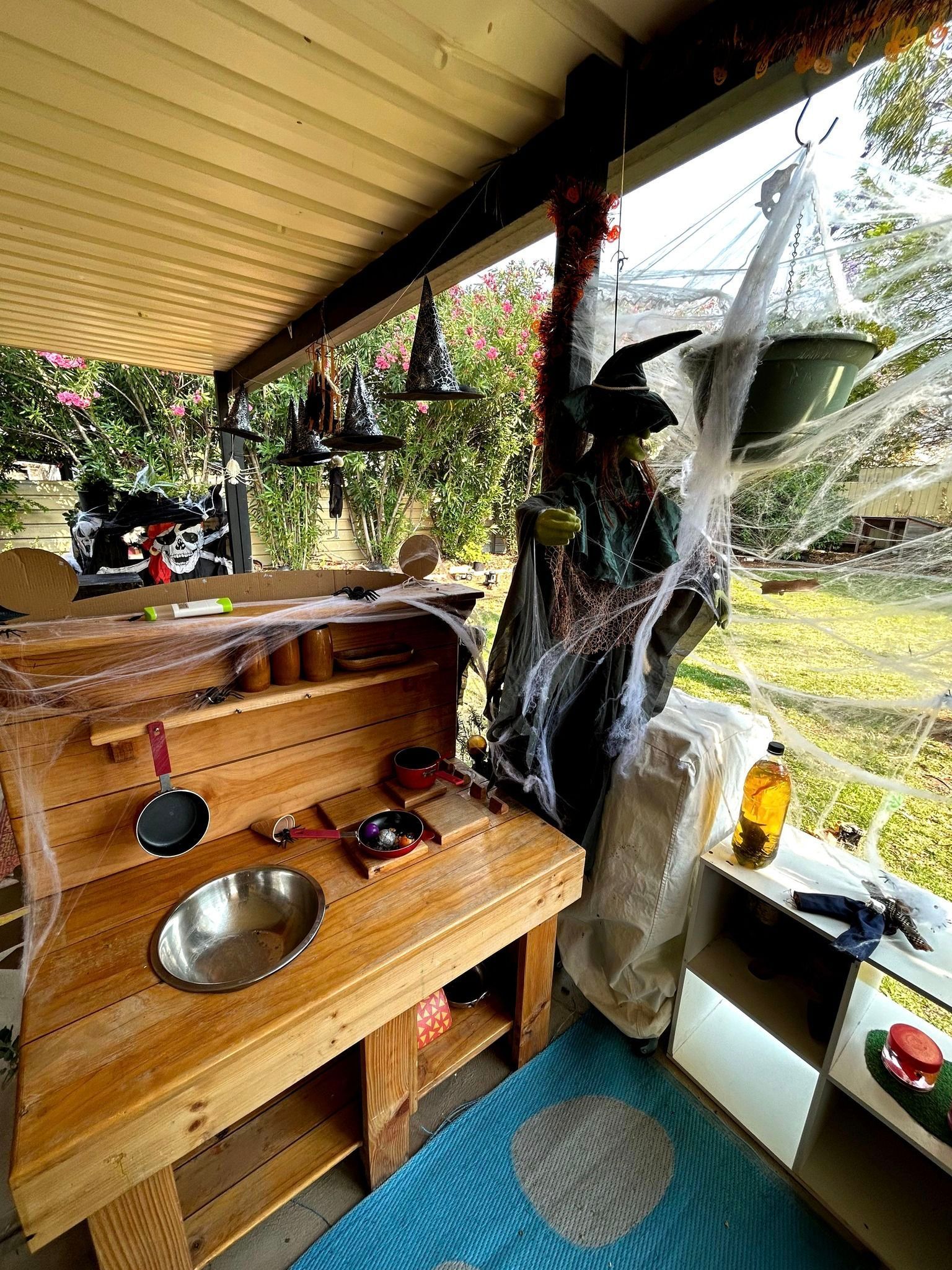 Halloween-decorated outdoor play kitchen with witch hats, spiderwebs, and green plants in a sunny yard.