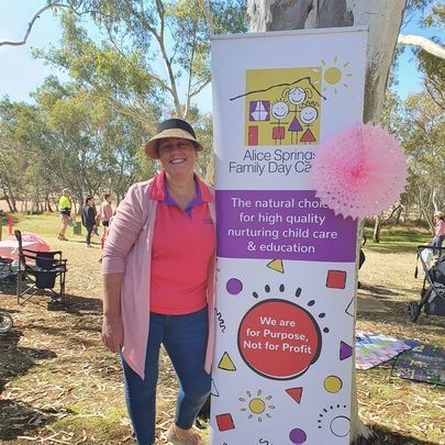 Woman in pink shirt and hat stands beside a banner for Alice Springs Family Day Care. Outdoor setting.