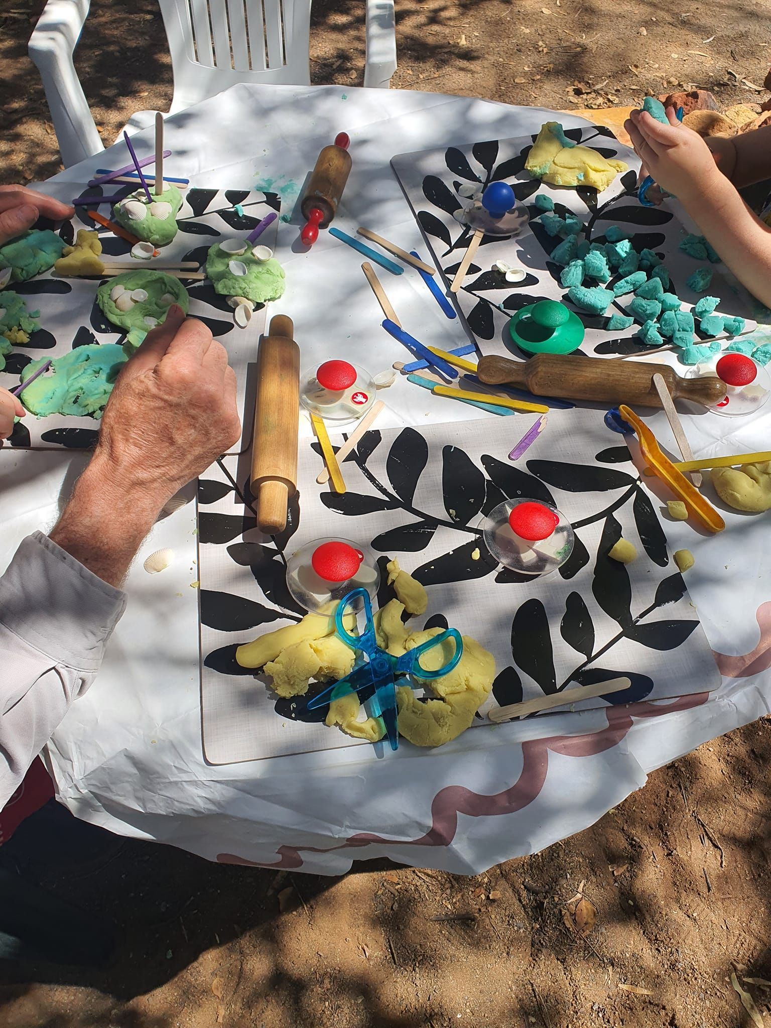 People sculpting with playdough at an outdoor table with rolling pins, beads, and decorative mats.
