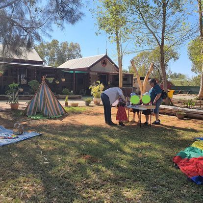 People and children at outdoor table, with teepee and buildings in background.
