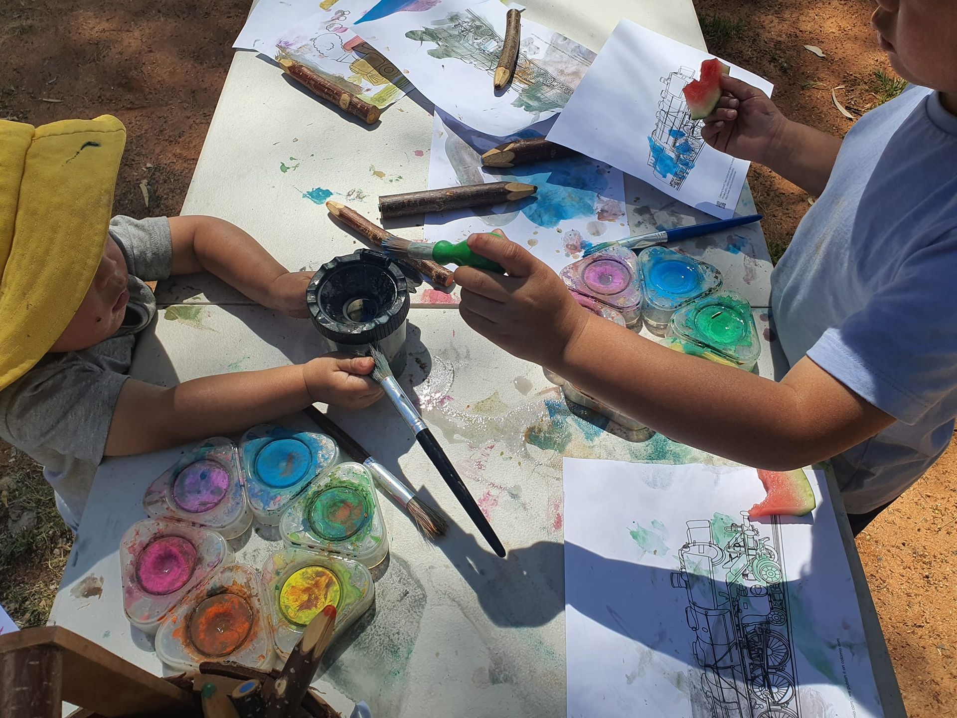 Two children painting with watercolors outdoors, using sticks and a rainbow palette.