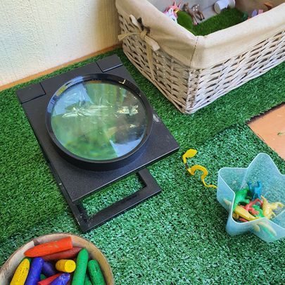 Magnifying glass on stand, looking at green carpet. Crayons, basket, toys nearby.