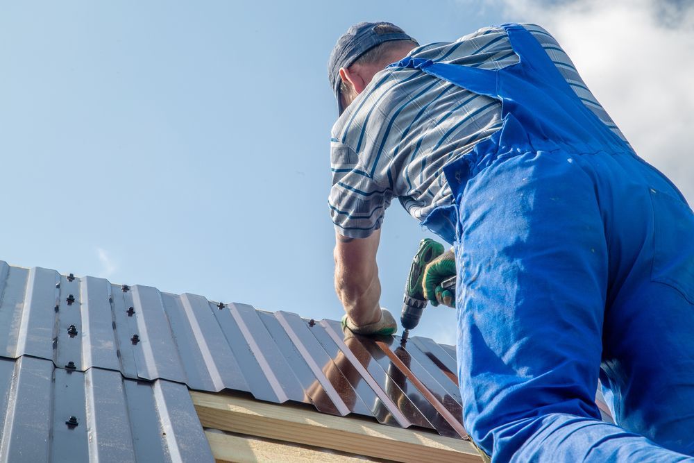 Roofer in Blue Overalls Drilling Into a Metal Roof Under a Blue Sky — Wilko Plate (Aust) Pty Ltd in Tully, QLD