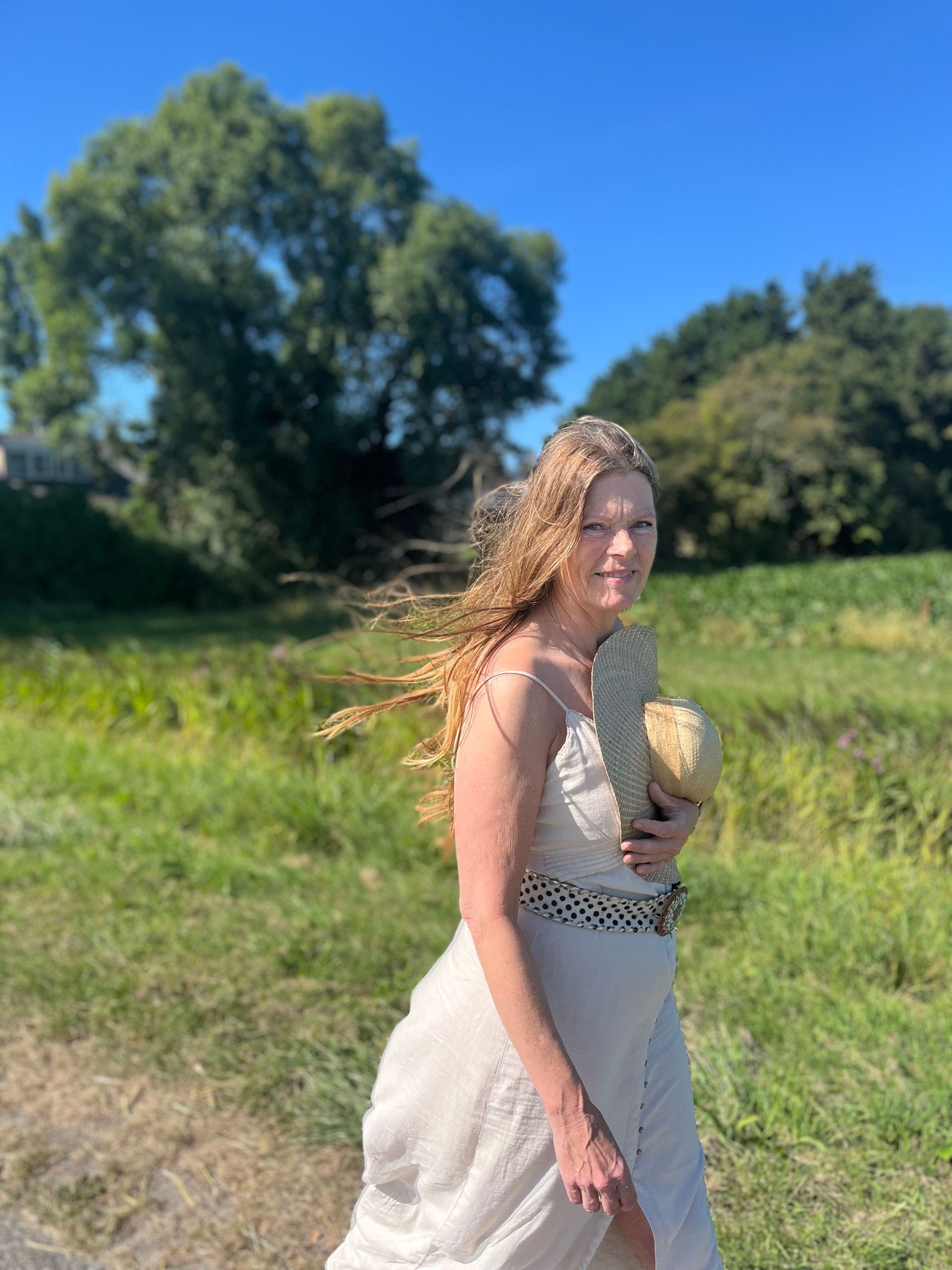 Vrouw in een witte jurk loopt in een zonnig grasveld met bomen en een blauwe lucht op de achtergrond.