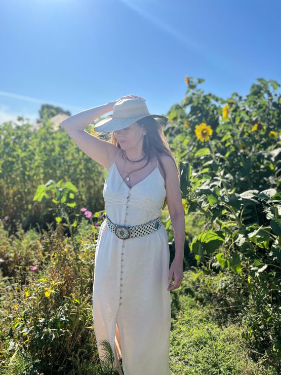 Een vrouw in een witte jurk en een strohoed staat in een zonnige tuin en houdt de rand van haar hoed vast.