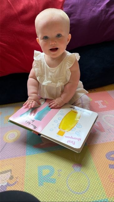 Baby doll in a cream dress sitting on a patterned rug, holding an open picture book.