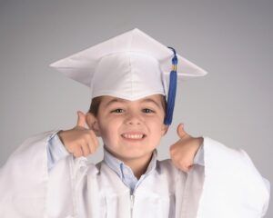 Smiling child in white graduation cap and gown giving two thumbs up against a gray background