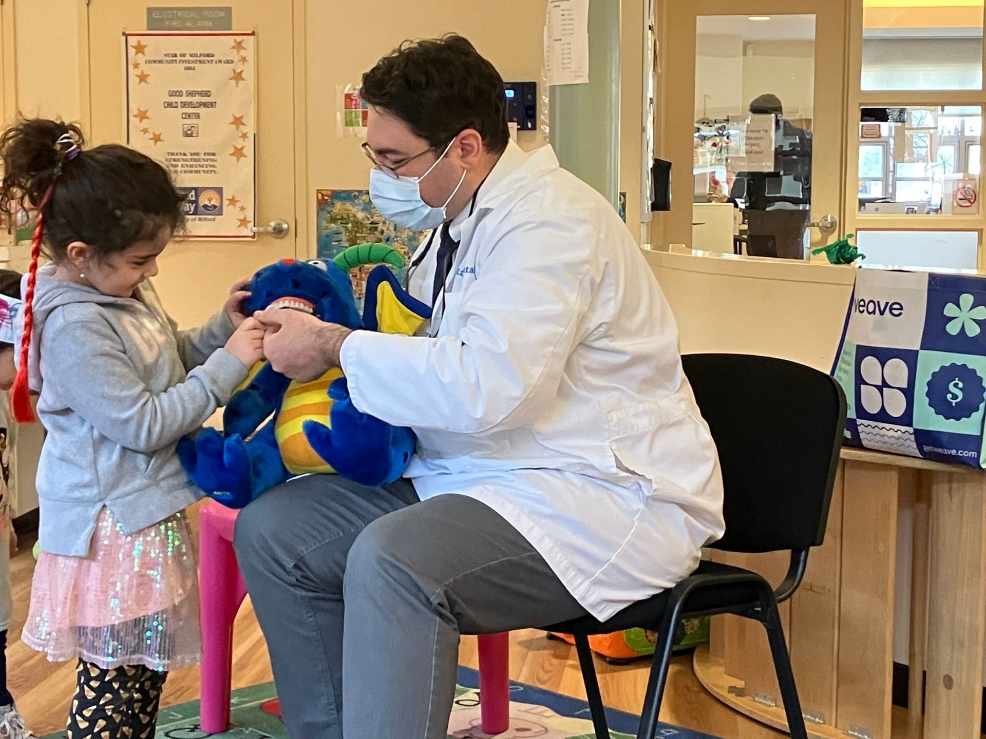 Doctor in white coat and mask helping a child hold a plush toy in a classroom.