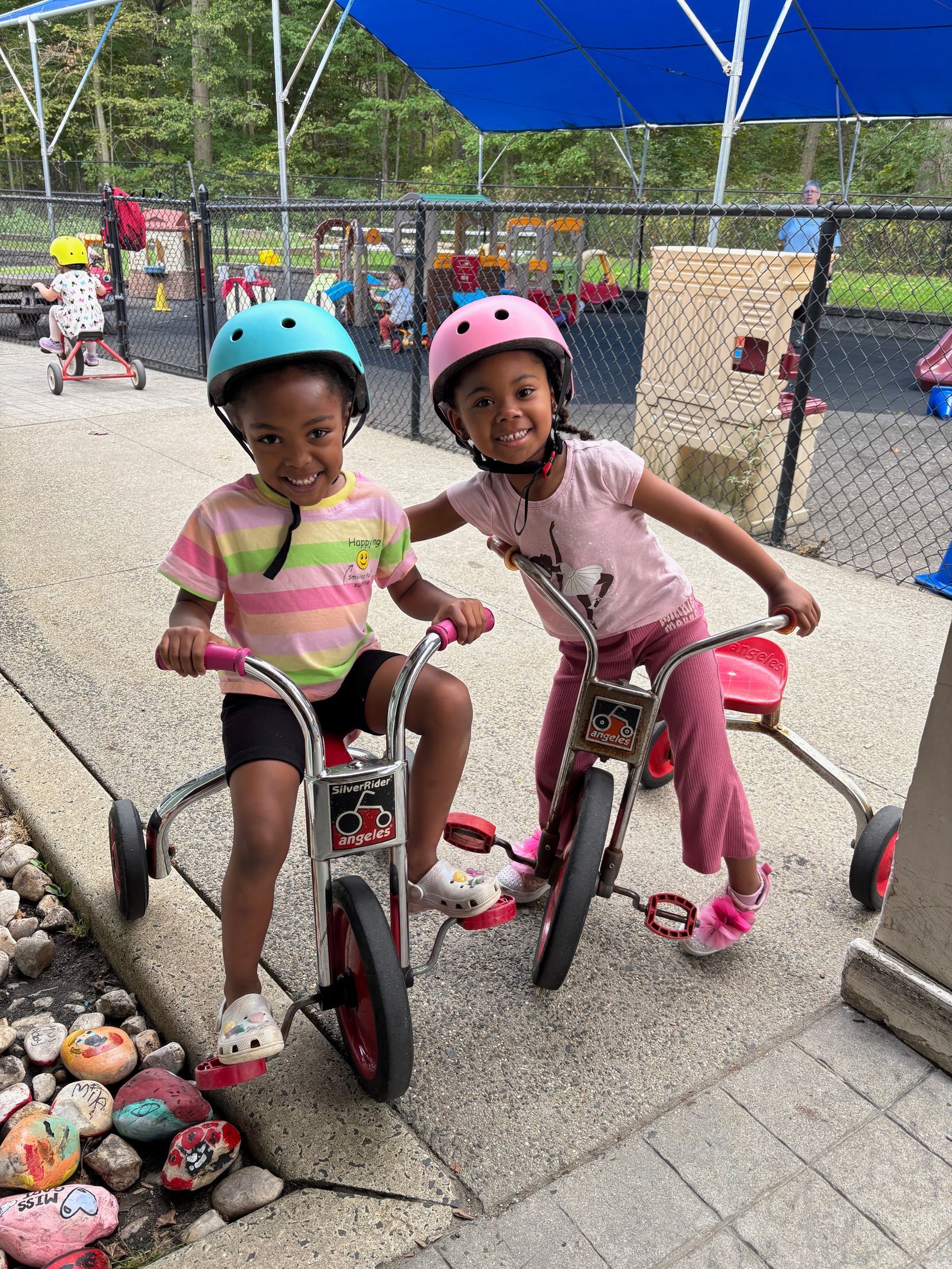 Two children wearing helmets pose on tricycles under a blue canopy outdoors