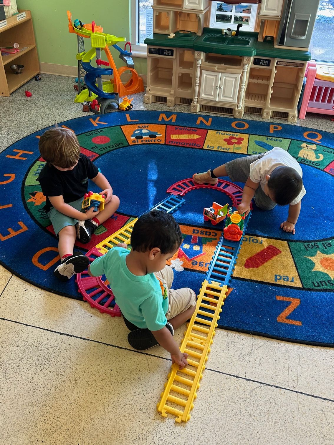 Children playing with toy tracks on a colorful classroom rug, building a winding road together.