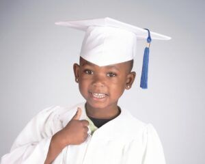 Smiling graduate in white cap and gown giving a thumbs-up against a plain background