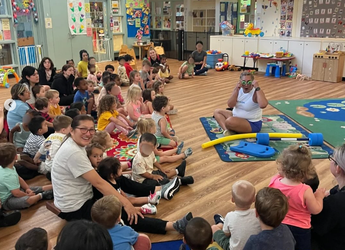 Doctor in white coat and mask helping a child hold a plush toy in a classroom.