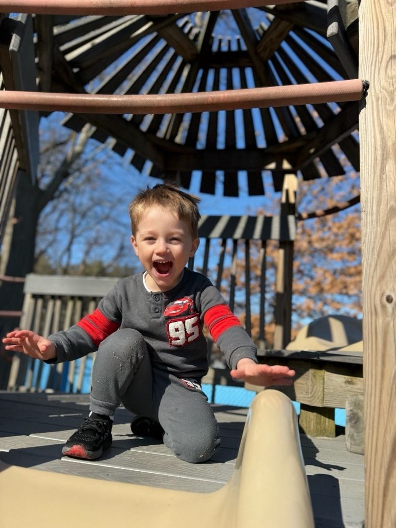 Smiling child kneels on a playground structure with arms outstretched under a wooden canopy.