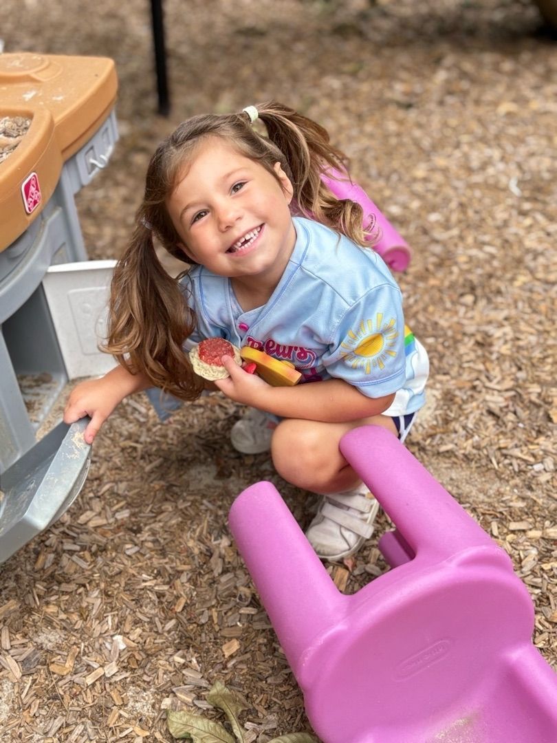 Smiling child crouches by a pink toy in a woodchip playground area