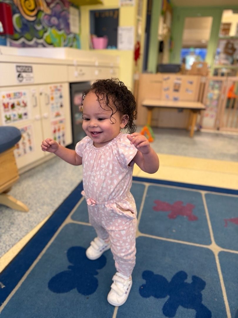 Smiling toddler standing on a blue rug in a colorful classroom, arms raised for balance