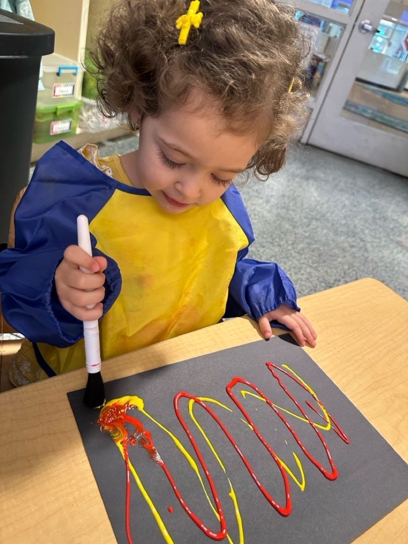 Child painting red and yellow wavy lines on black paper at a table