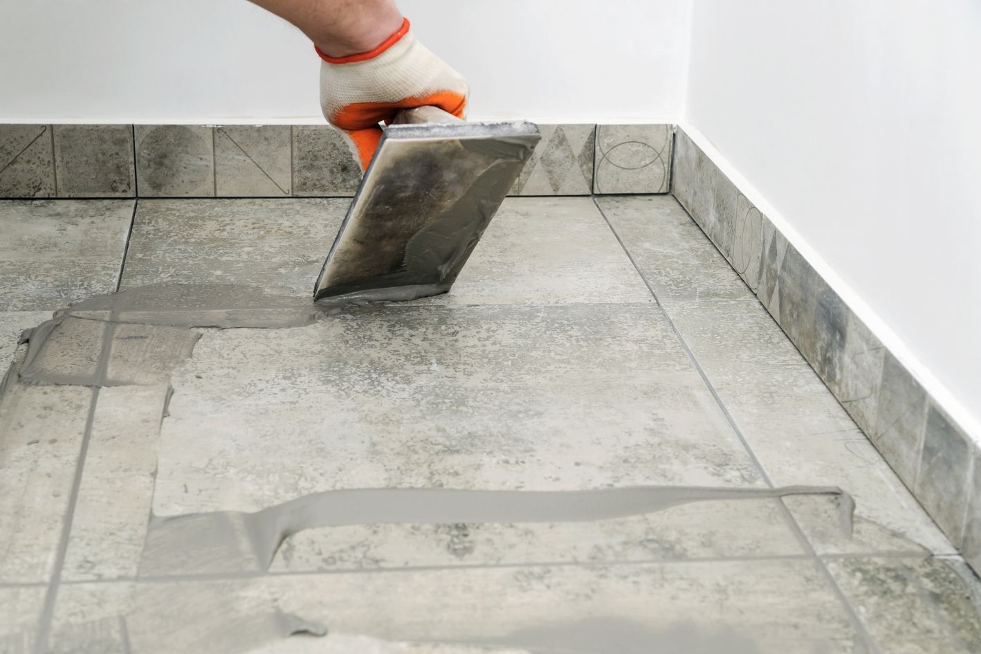 Person using a trowel to apply gray grout to tile flooring in a corner of a room.
