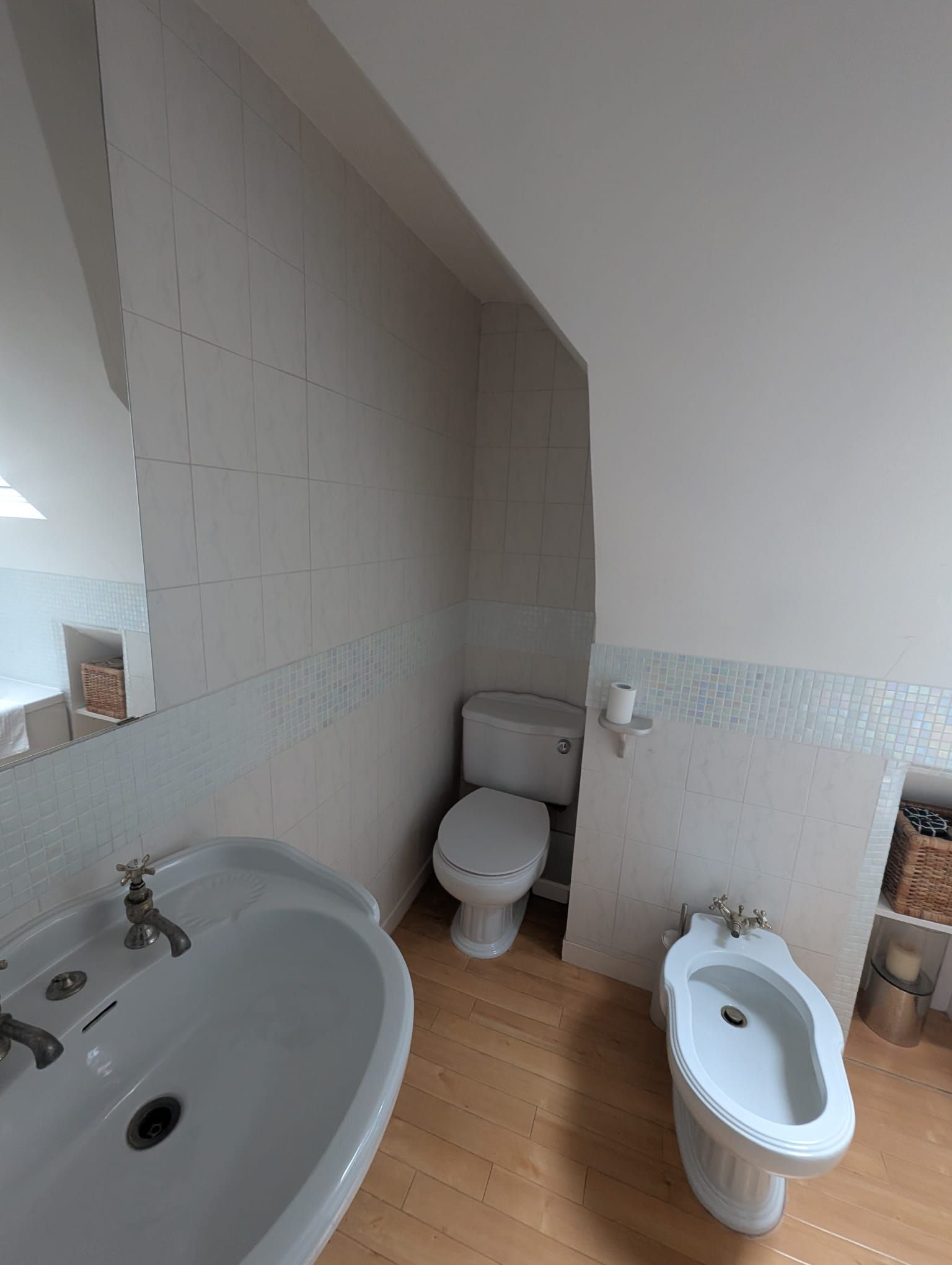 Bathroom with toilet, bidet, sink, and mirror. White tile and walls with wood flooring.