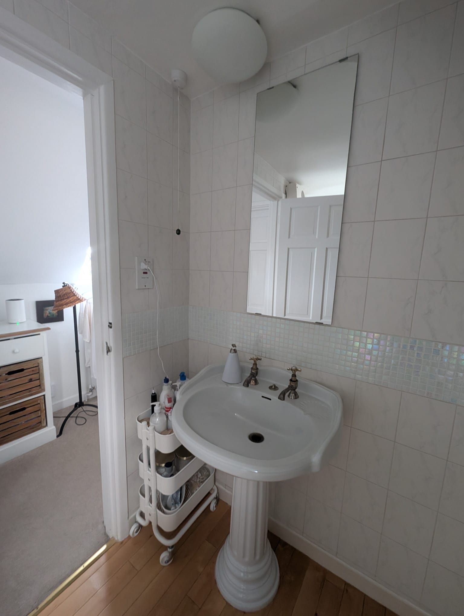 White bathroom with pedestal sink, mirror, and rolling storage cart. Doorway to a bedroom.