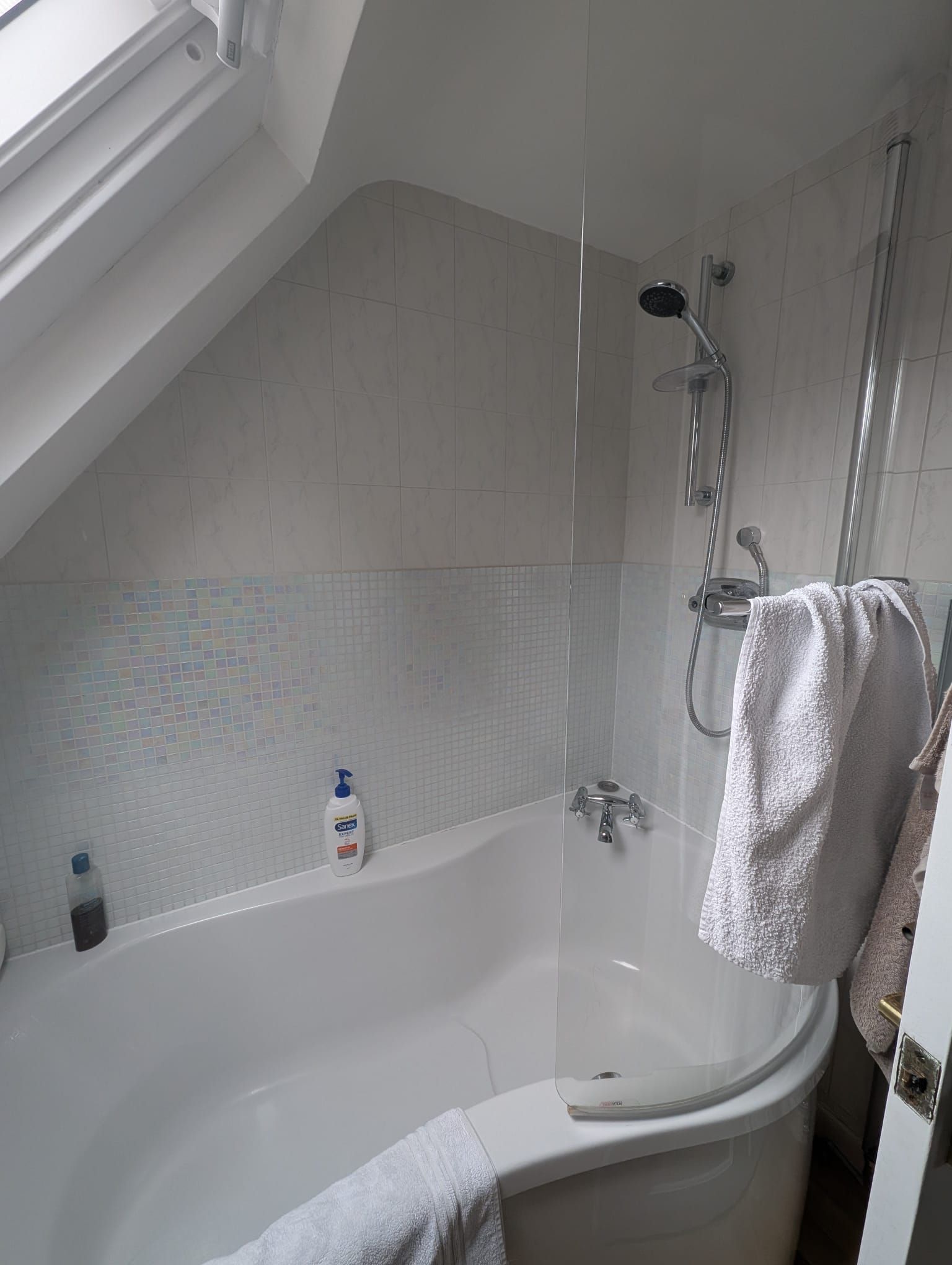 White bathroom with bathtub, shower, and slanted ceiling. Towels hang near the shower.
