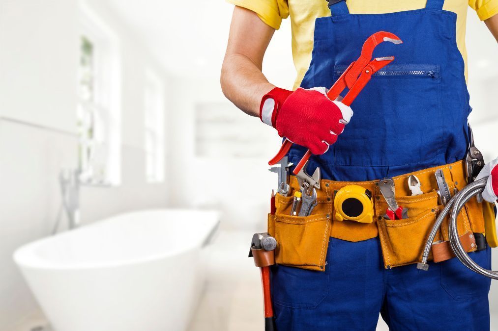 Plumber in blue overalls, holding wrench, tool belt with various tools, standing in a bathroom.