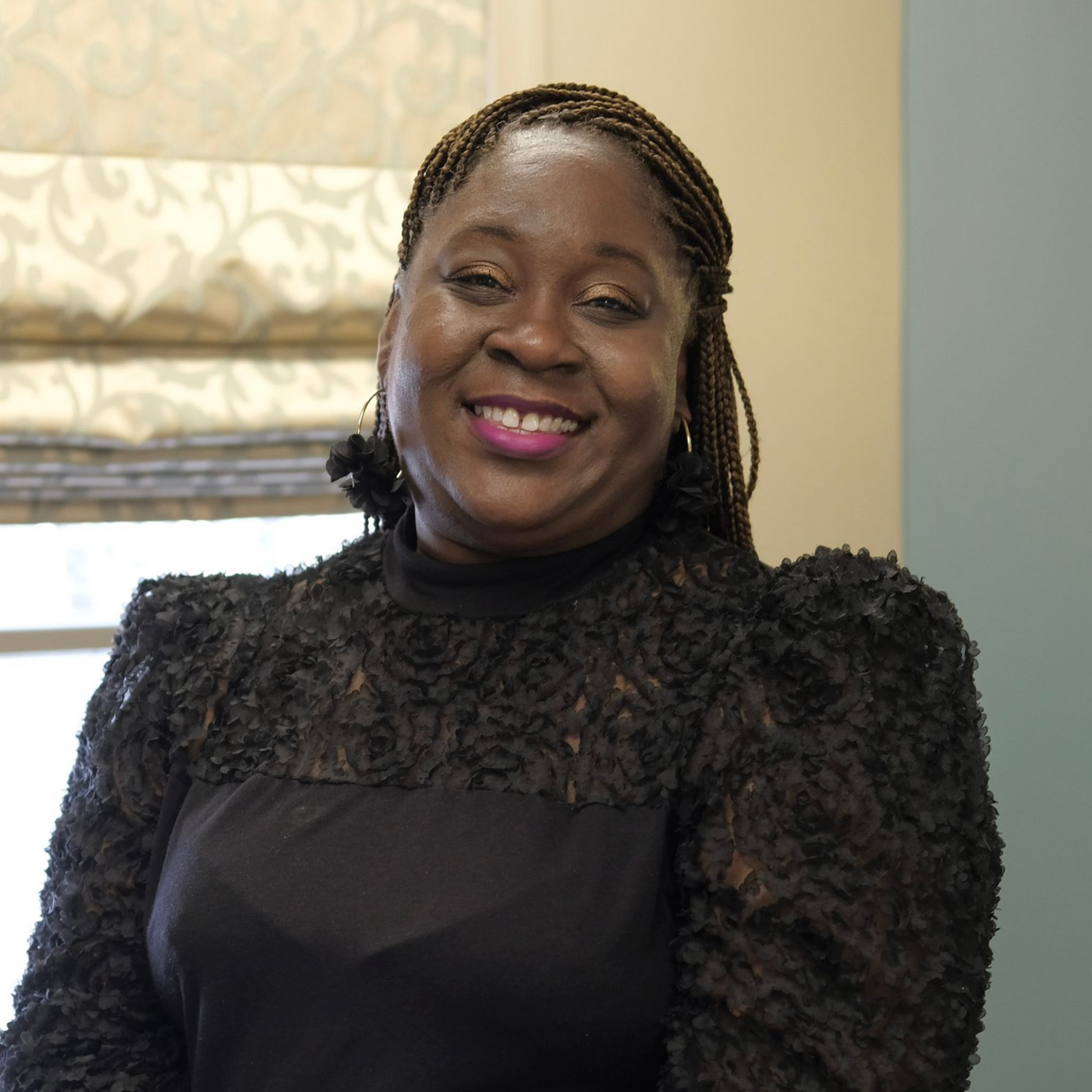 Gina with dreadlocks, wearing a black jacket and hoop earrings, smiling against a light blue background.