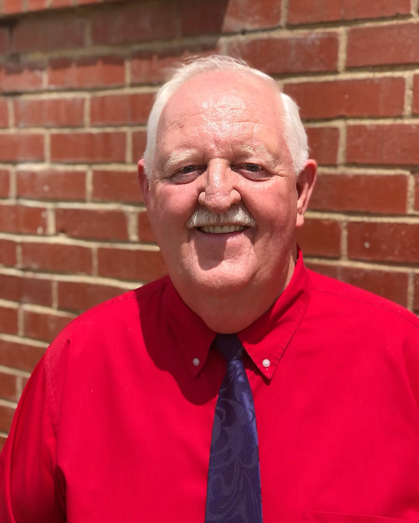 A man in a red shirt and blue tie is smiling in front of a brick wall.