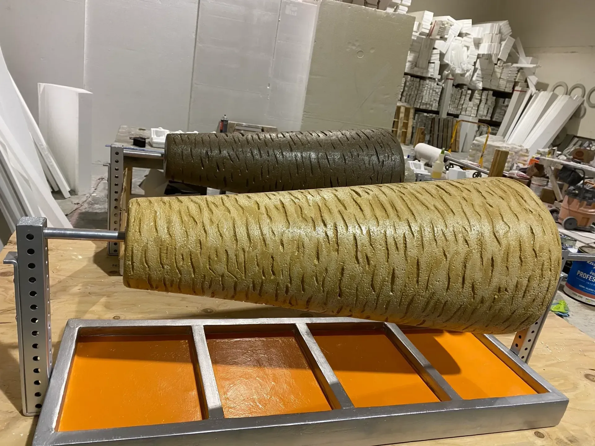 Two large textured rolls on a metal rack above orange containers in an industrial workshop