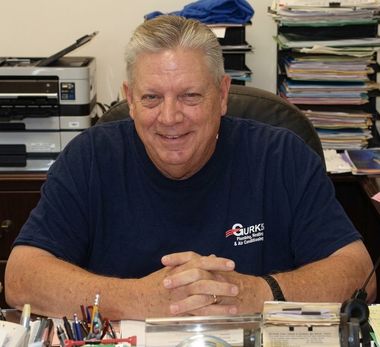 Man sitting at an office desk, smiling with hands clasped, surrounded by papers and office supplies.