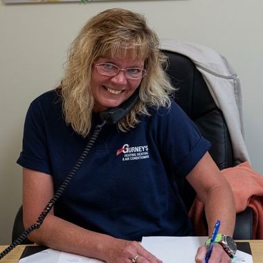 Smiling receptionist at desk, wearing glasses and navy shirt, speaking on a phone and writing notes.