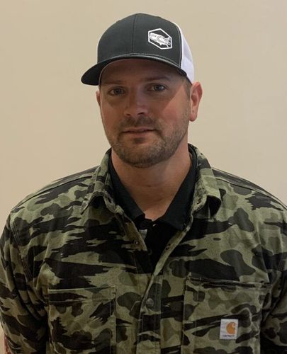 A man wearing a hat and a camouflage shirt is standing in front of a wall.