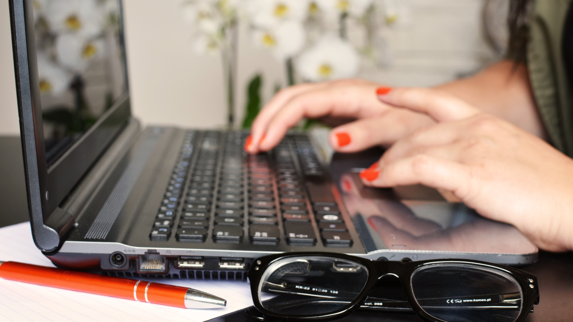 A woman is typing on a laptop next to a pair of glasses and a pen