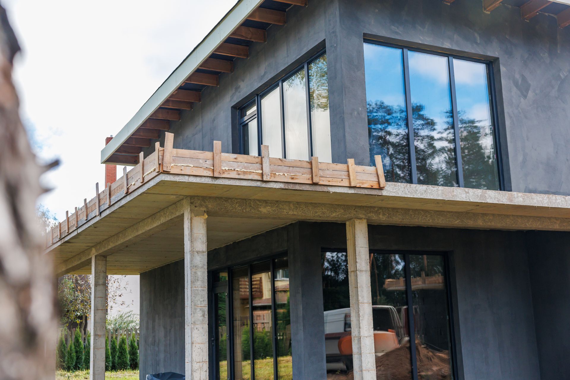Modern two-story house under construction with concrete columns, large windows, and a wooden balcony.