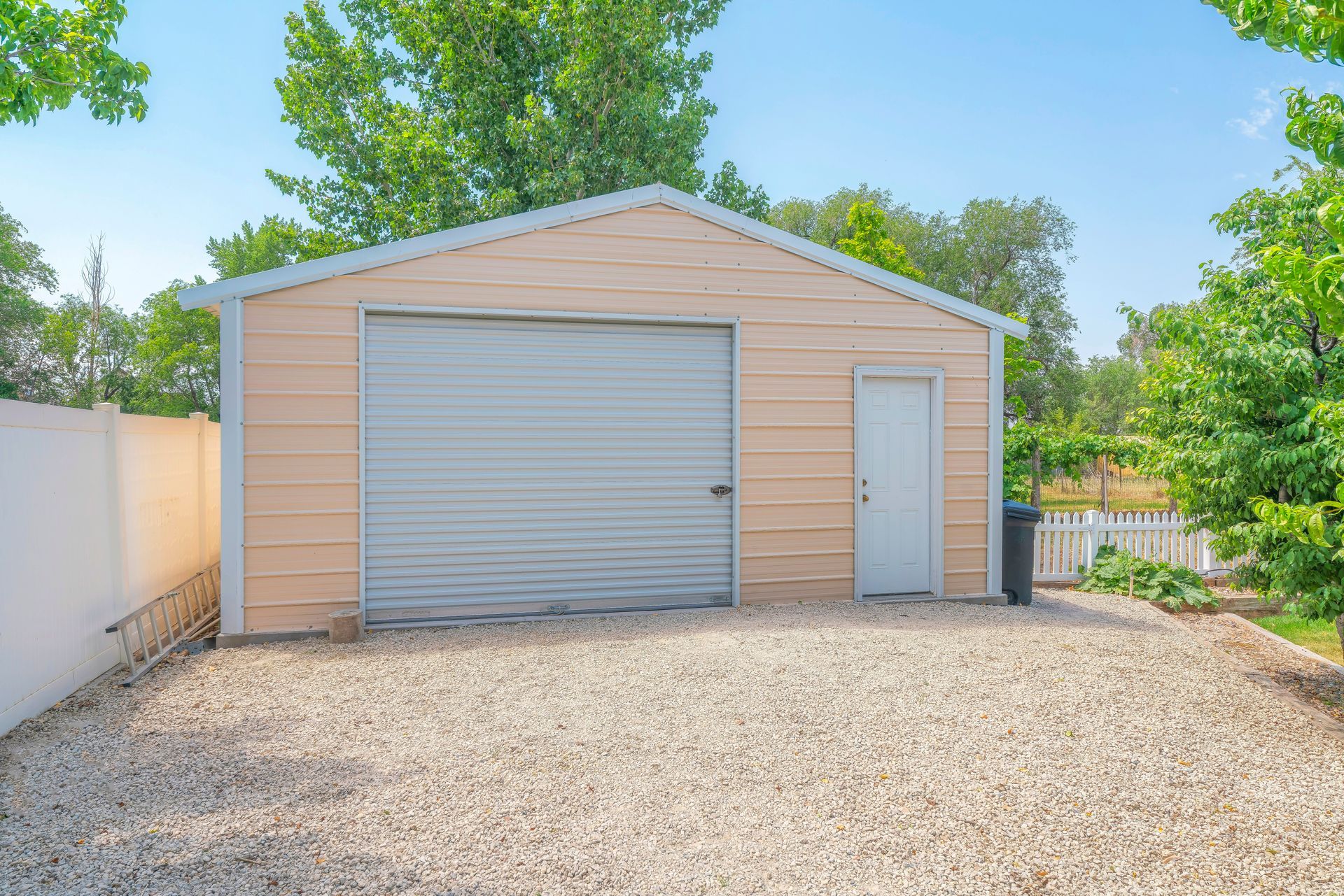 Beige detached garage with a large roll-up door and side entrance on a gravel driveway under a blue sky