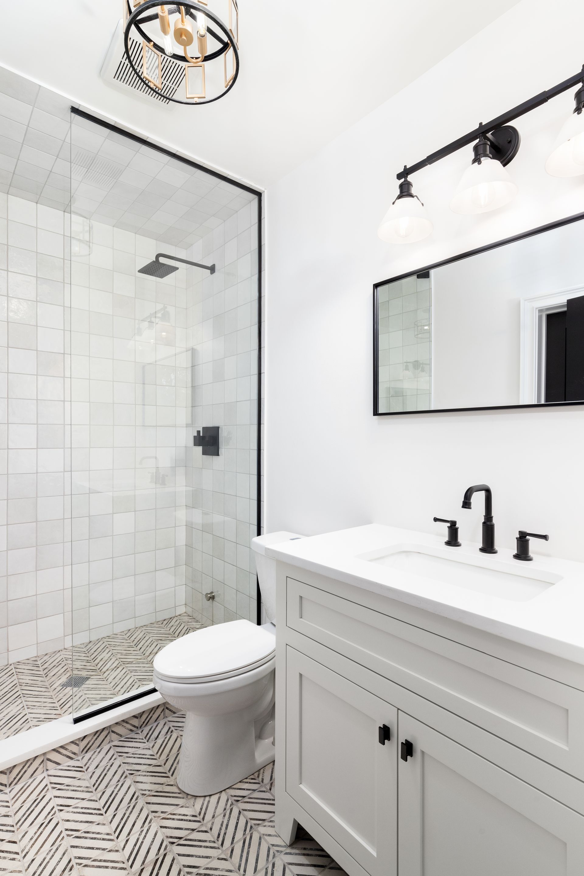 Modern white bathroom with herringbone floor tile, glass shower, and black fixtures.