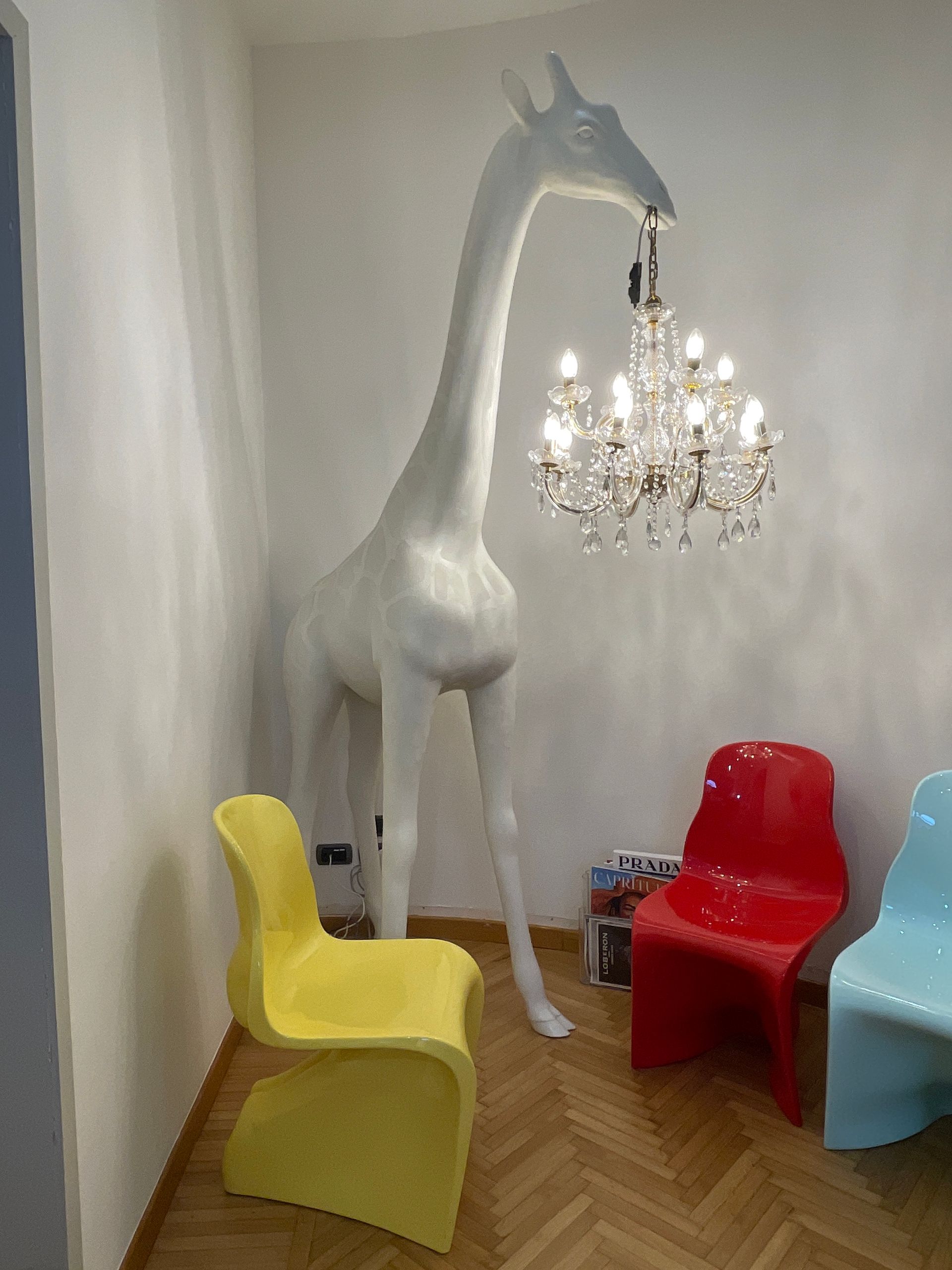 Scultura di giraffa bianca che regge un lampadario; sedie colorate in un angolo.