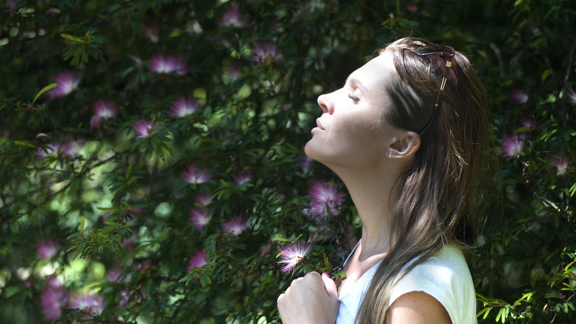 A woman is standing in front of a bush with her eyes closed.
