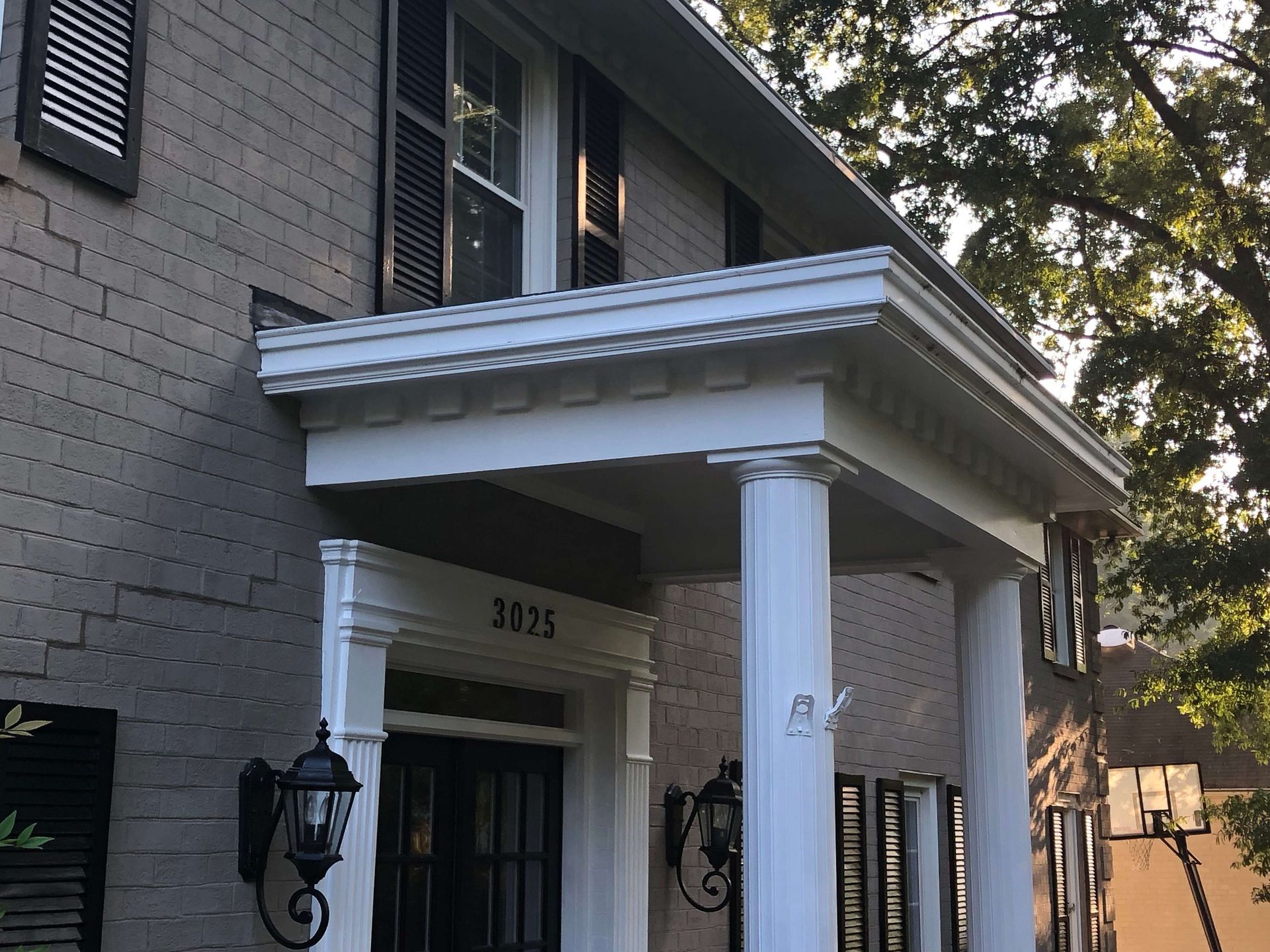 Two-story house with columns and a covered porch. Gray brick exterior, black shutters, white trim, and a basketball hoop.