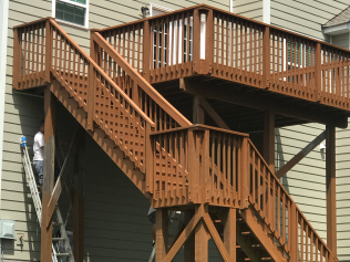 Brown wooden deck with stairs attached to a light-colored house; person on a ladder painting.