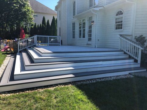 Multi-level gray deck with white railings and stairs, attached to a two-story white house, sunny day.