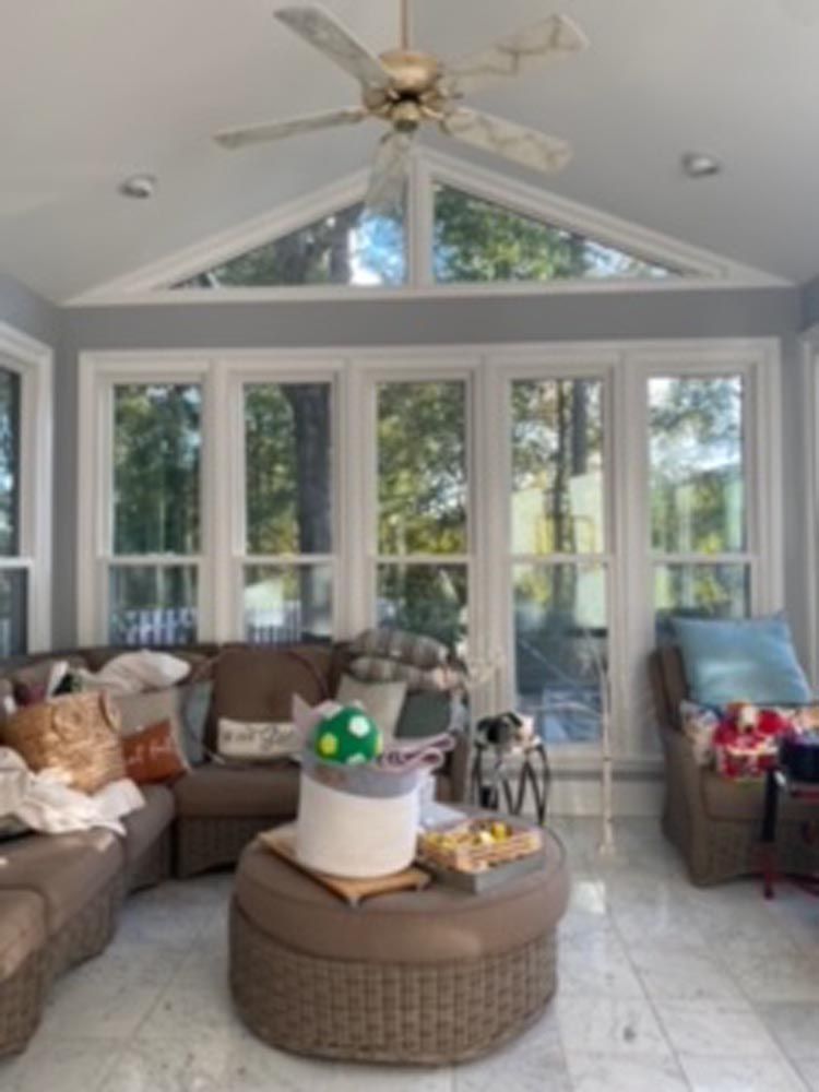 Sunroom with sectional sofa, round ottoman, and large windows looking out to trees.