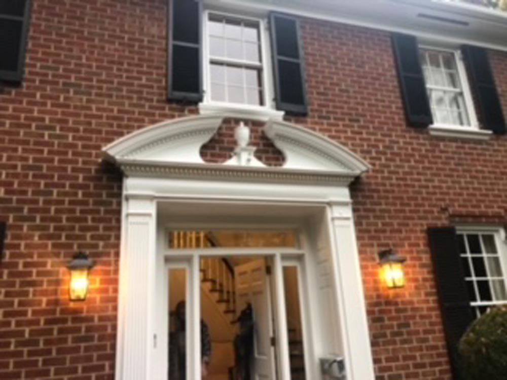 Red brick house with white trim around the door, black shutters, and two sconces with lit orange light.