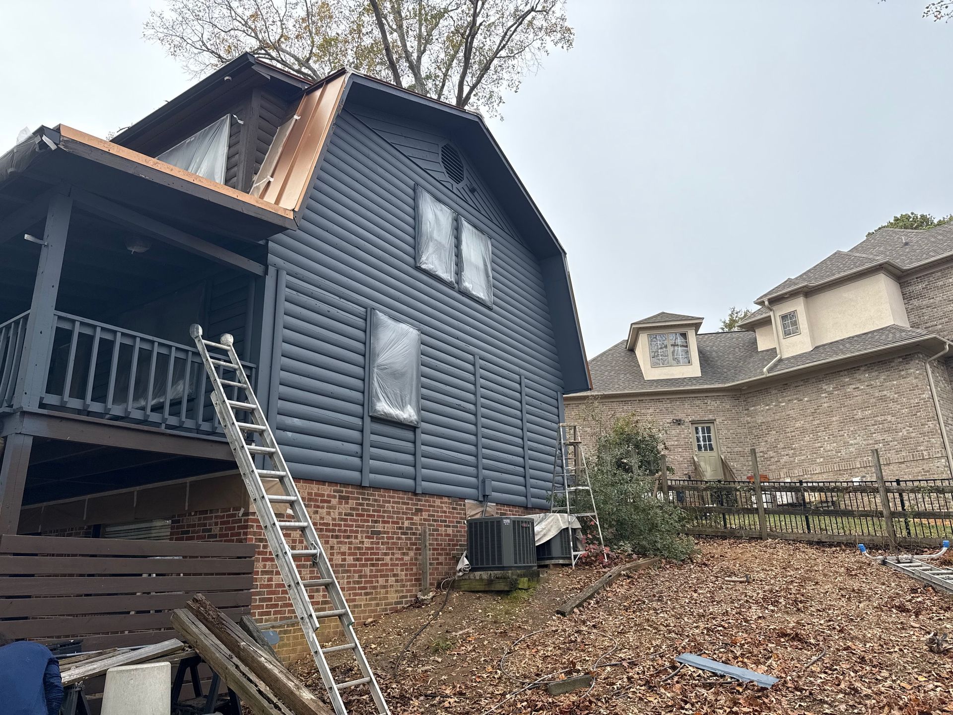 Dark blue house with ladder, brick foundation, and nearby trees, under cloudy sky.