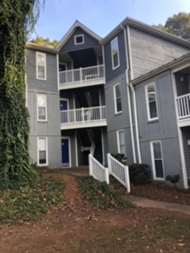 Three-story gray apartment building with white balconies and stairs. Dark blue door and ivy on the left.