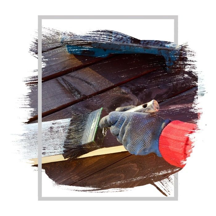 Person with gloved hand painting wooden planks with a brush; dark stain applied.