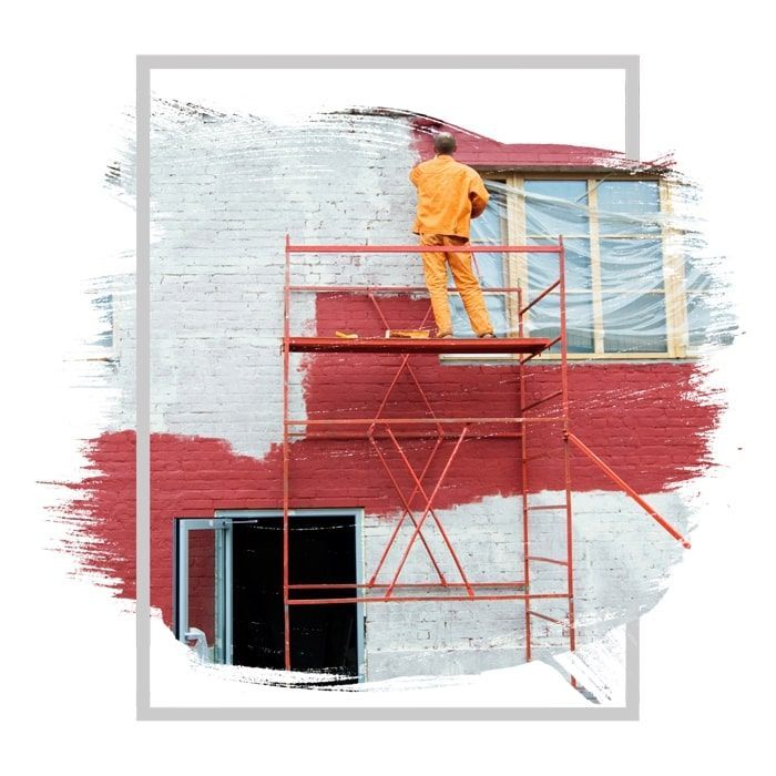 Person painting a red brick wall white, standing on a scaffold outdoors.