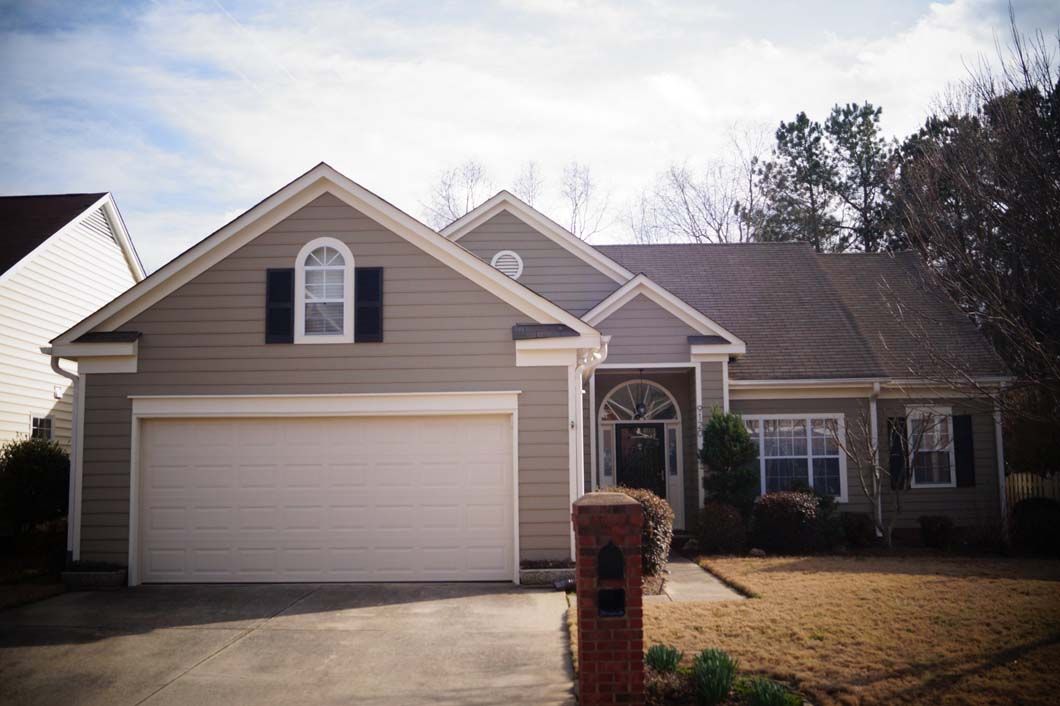 Tan house with white garage door, black shutters, brick mailbox, brown roof, and small front yard.